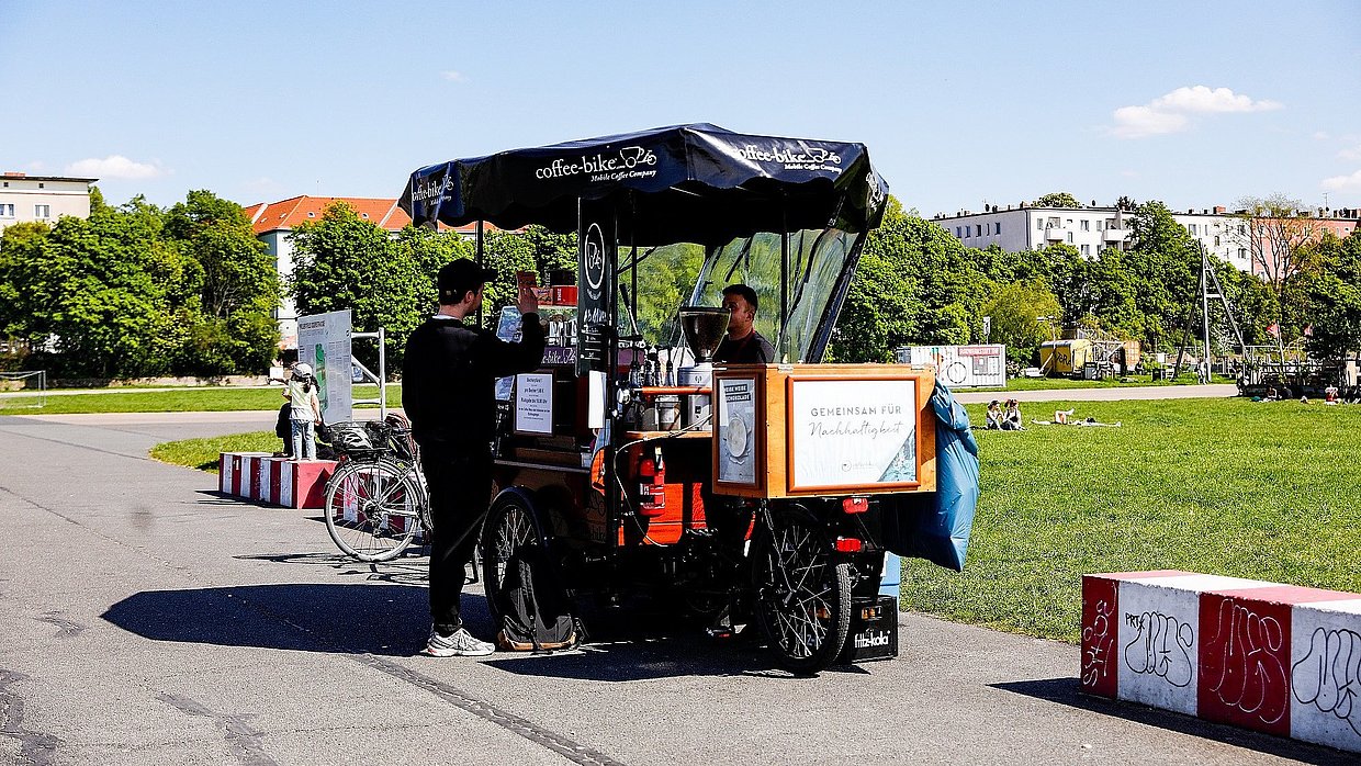Ein in schwarz gekleideter Mensch steht vor einem Coffee-Bike auf dem Tempelhofer Feld
